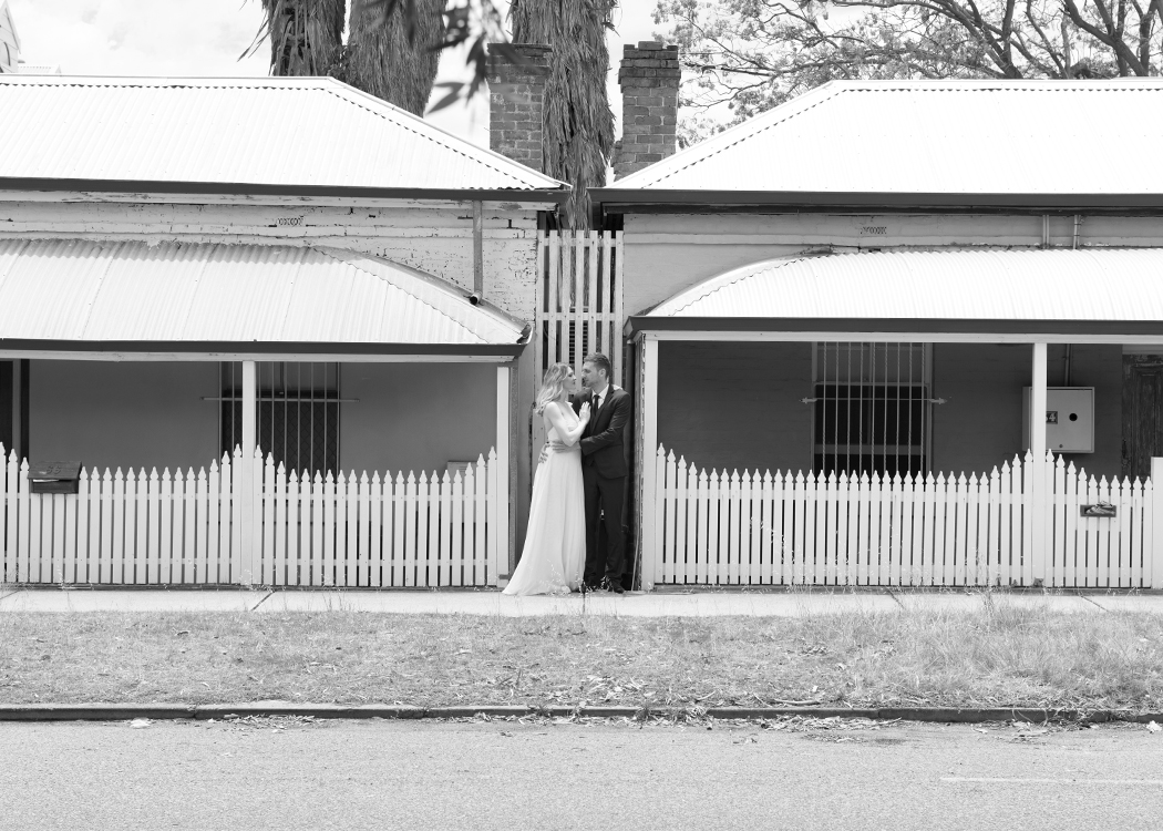 Bride and groom standing between two old houses with white picket fence