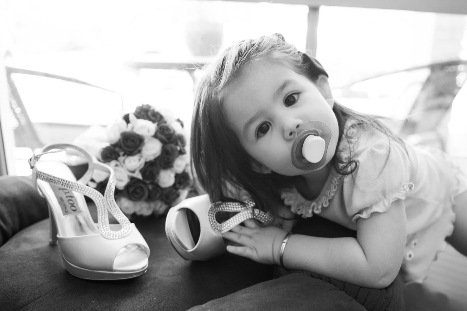 Black and white photo of little girl with dummy and wedding shoes and bouquet. Gazing at the camera. Wedding toddler