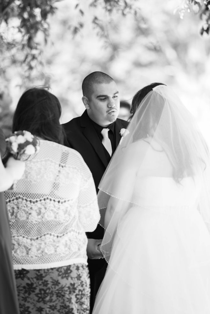 Black and white photo of groom during wedding ceremony, tender moment. Looking at his bride holding hands