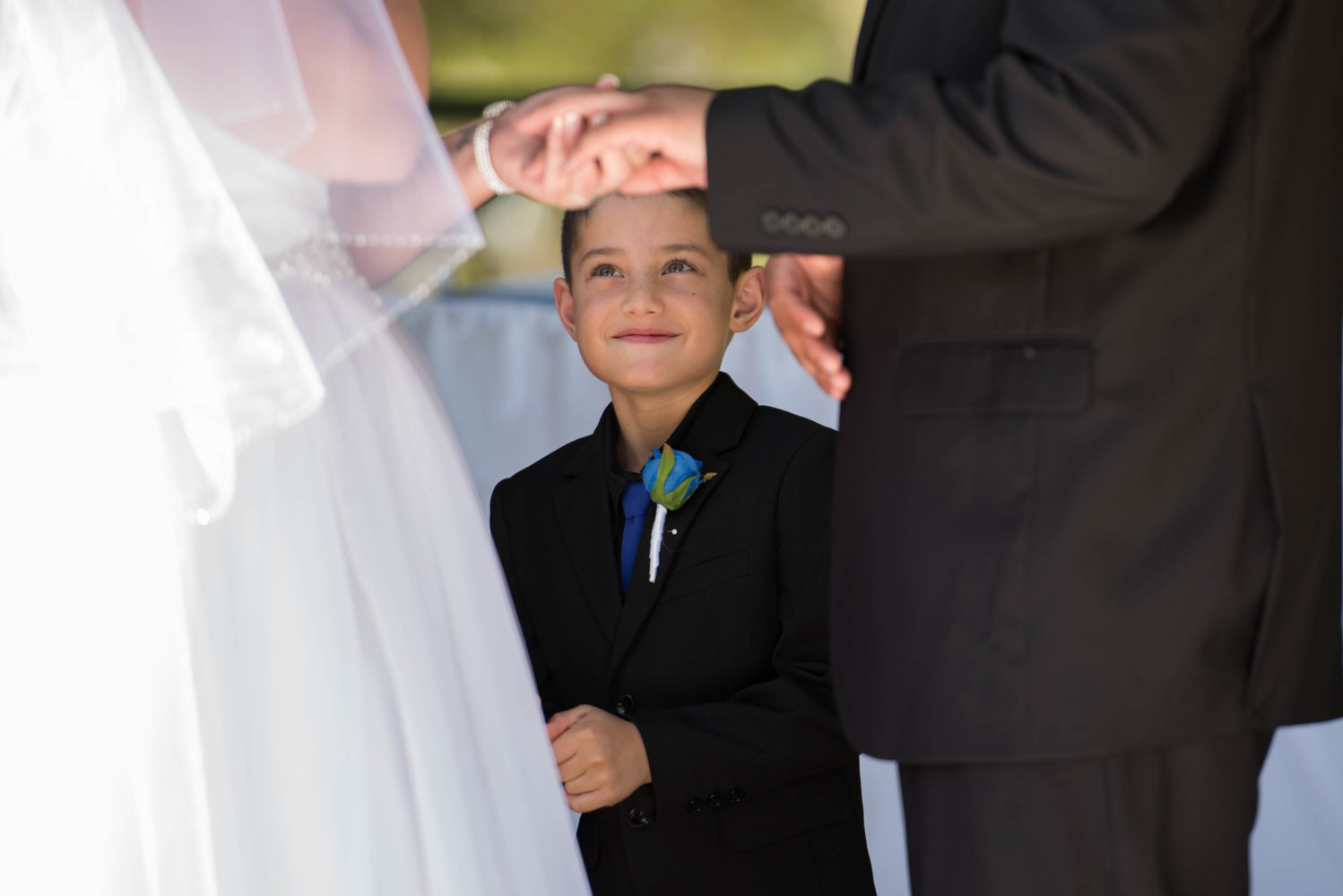 Son smiling and beaming at his mum as she exchanges rings during the wedding ceremony. Blue flower buttonhole