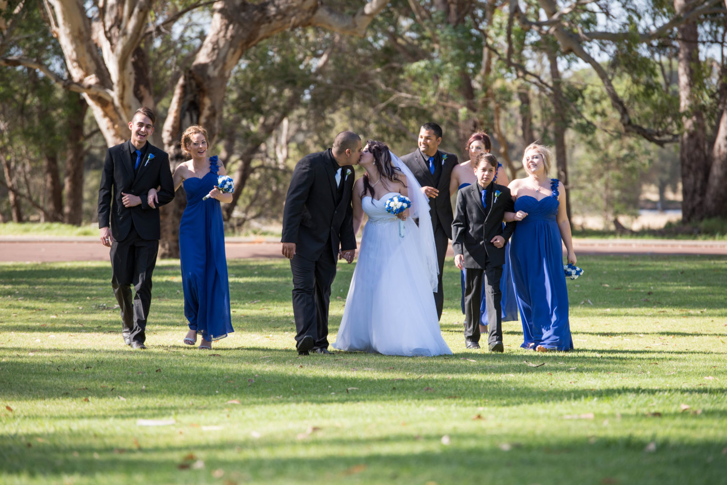 Colour candid photo of bridal party walking towards the camera at a distance. Bride and groom kissing.