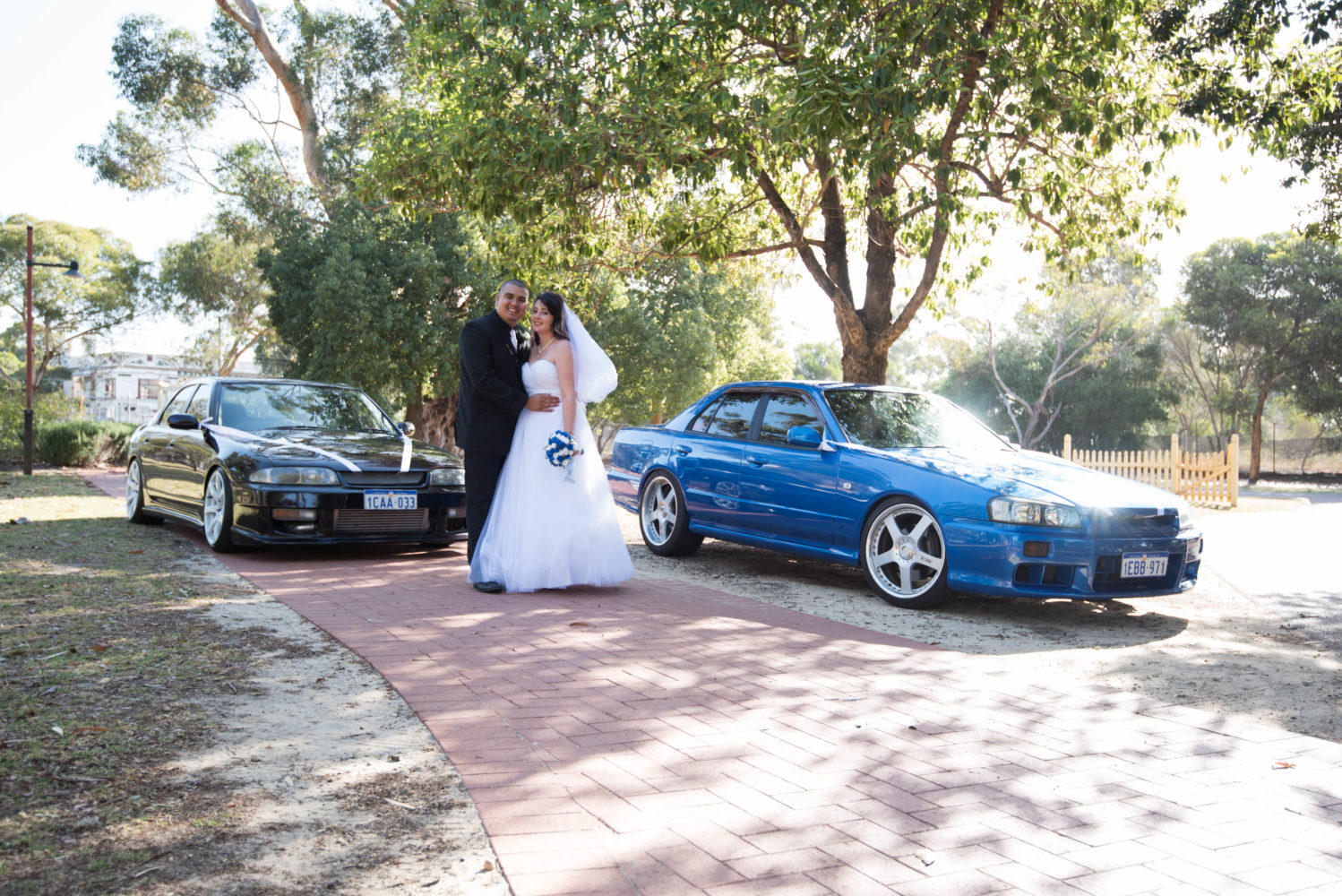 Bride and groom standing with their blue and black cars. Wedding ribbon on cars. Muscle cars.