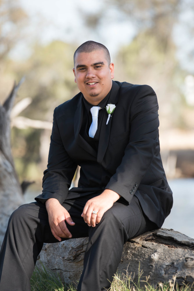 Colour midshot photo of groom smiling at the camera. Black suit white tie and white buttonhole