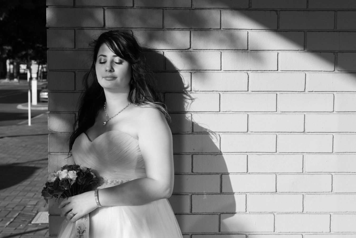 Black and white photo of bride leaning against a white brick wall. Eyes closed