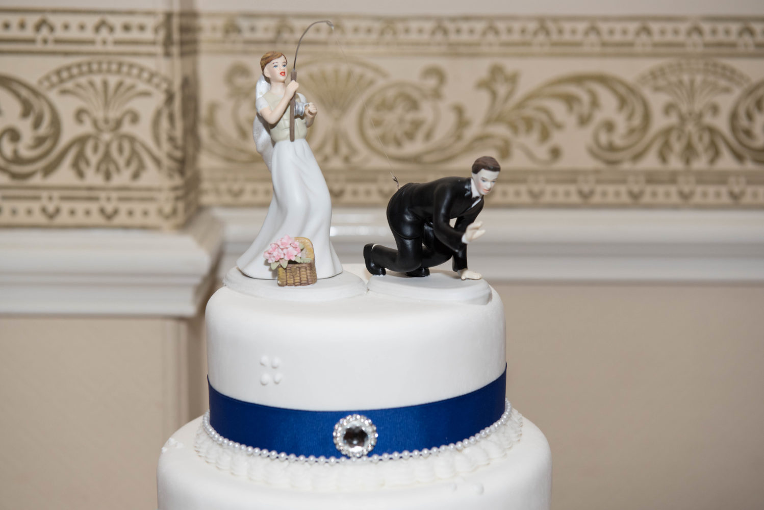 Close up photo of wedding cake toppers. Bride fishing for her husband. His pants are hooked by the fishing hook