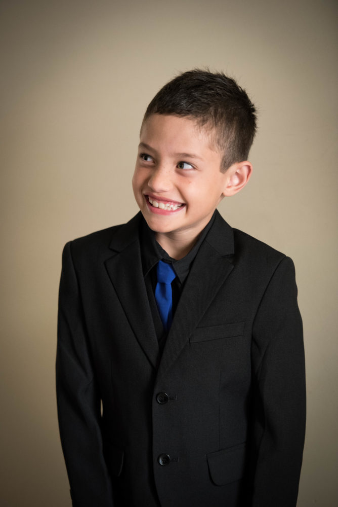 Colour photo of cheeky and happy looking boy on his dad's wedding day. Looking away from camera. Black suit blue tie