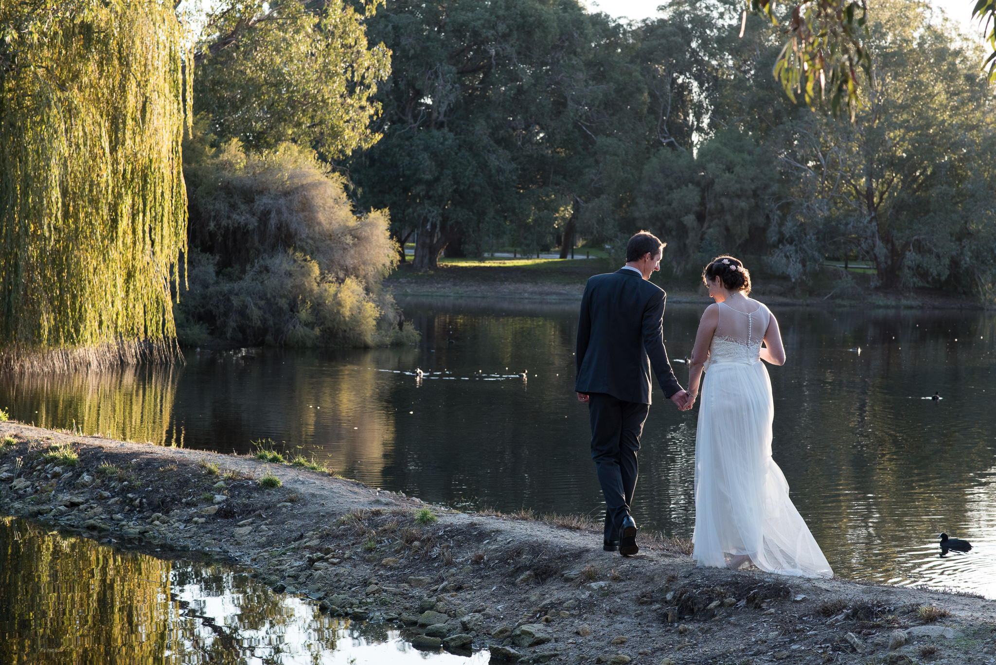 bride and groom looking down as they walk by a lake with weeping willow trees