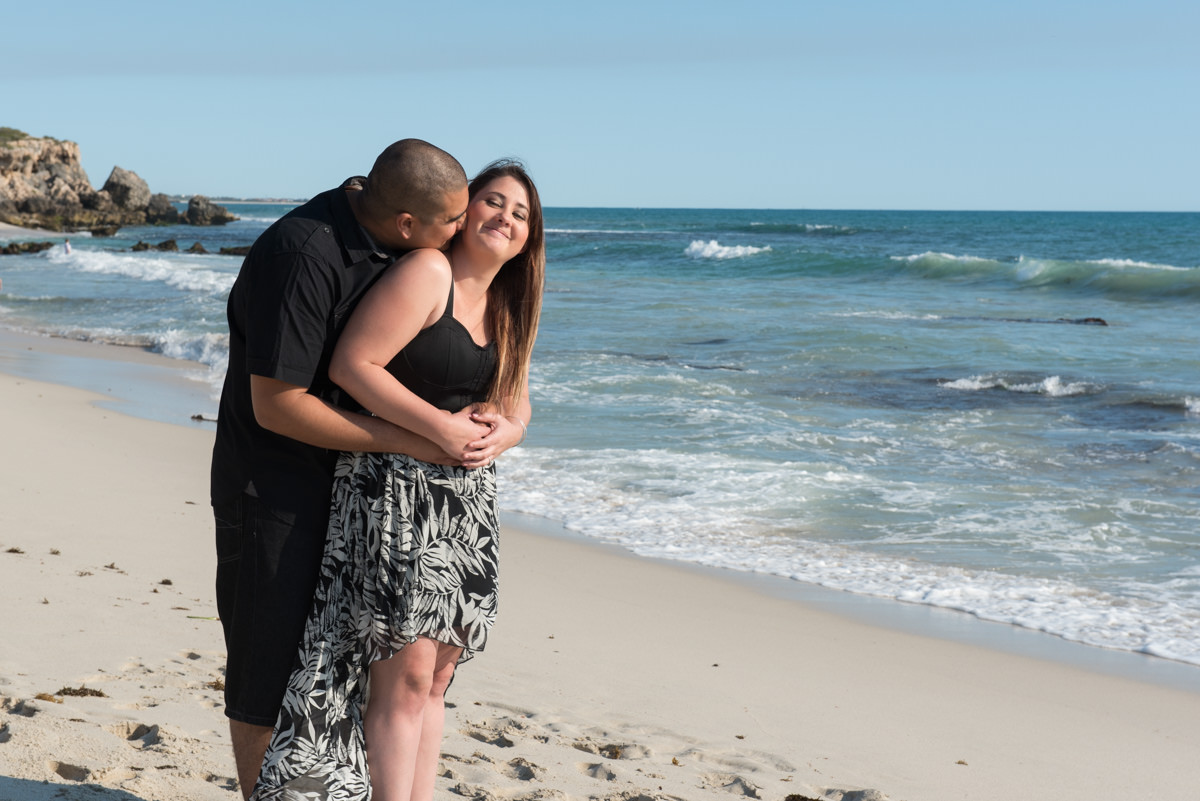 guy kisses his fiance's neck on the beach