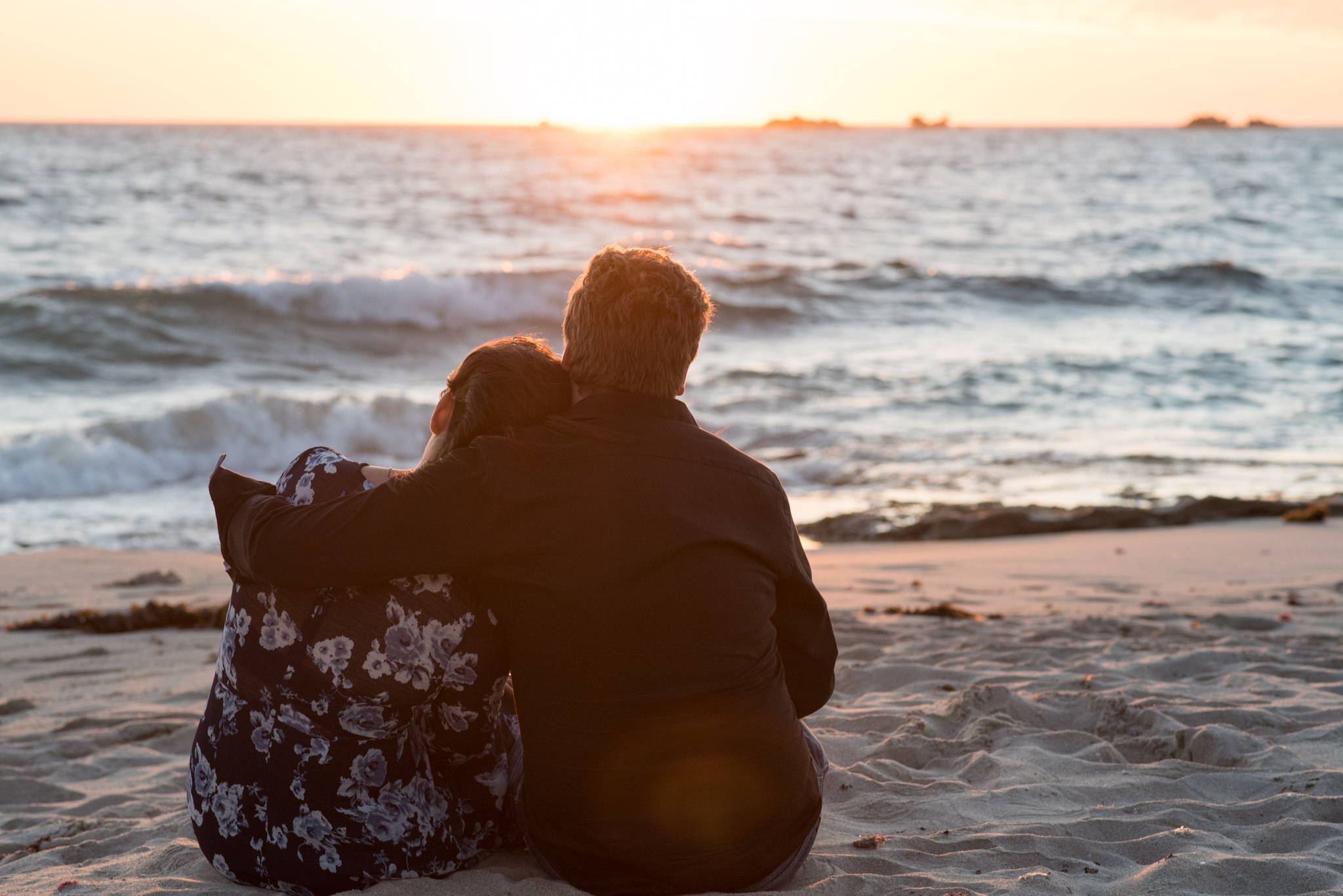 Man with his arm around girl while they watch the sun set on the beach