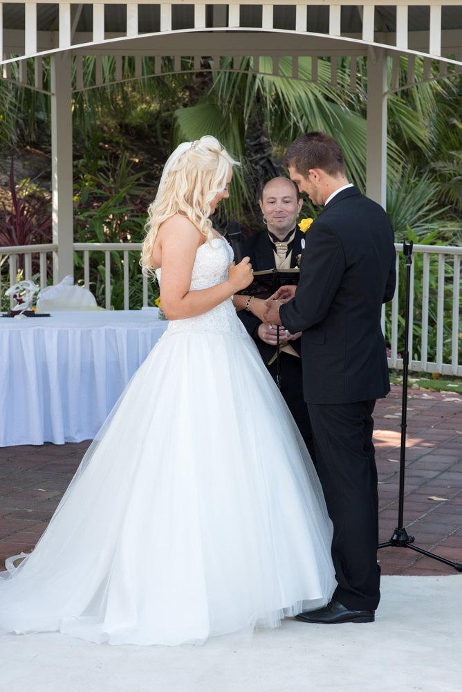 Groom puts ring on brides hand with celebrant behind