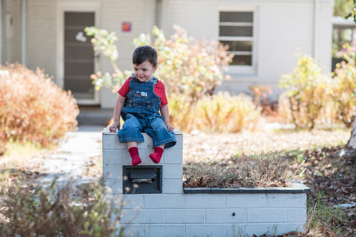 Little boy sitting on top of old letter box wearing red socks, red t-shirt and blue denim overalls