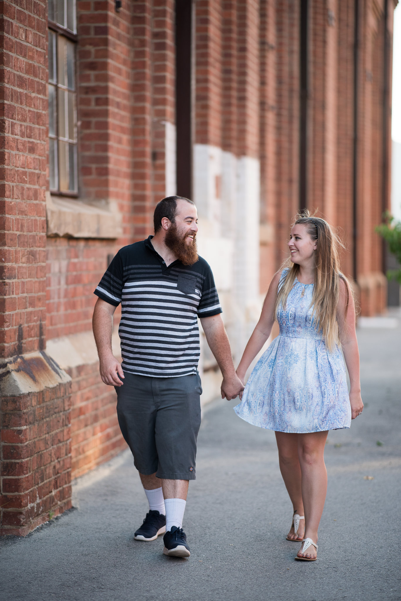 engaged couple smiling and walking along holding hands in front of red brick buildings