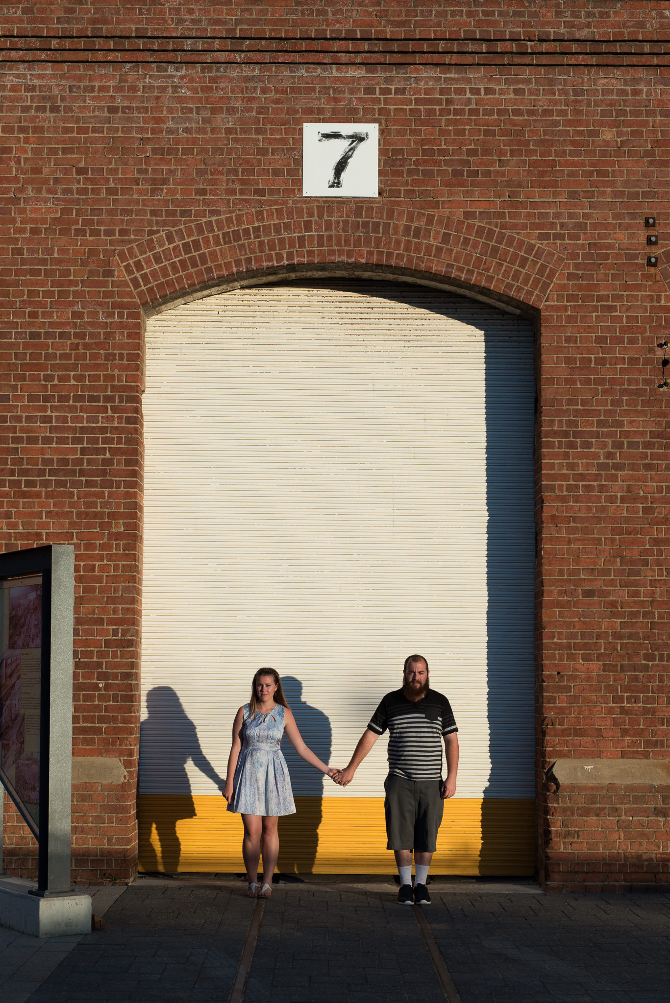 engaged couple standing apart holding hands with serious face infront of yellow and white shed door