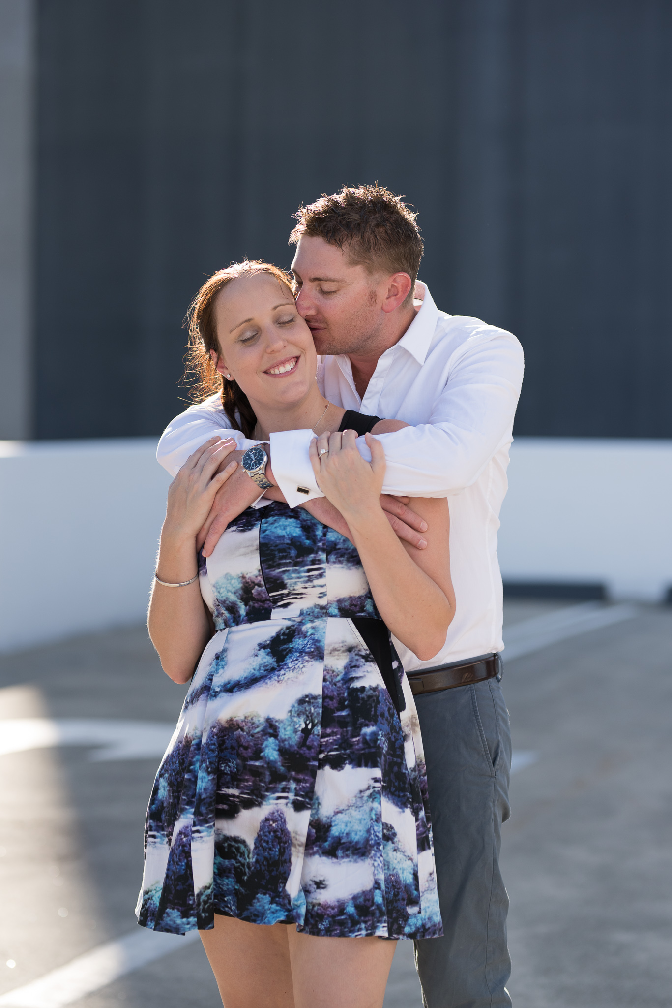 Boy hugs his fiancee on carpark rooftop with late afternoon sunlight