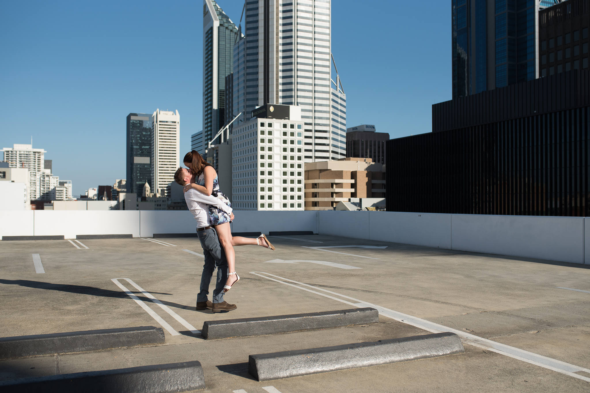 Boy lifts his fiancee up on carpark rooftop with Perth cityscape behind