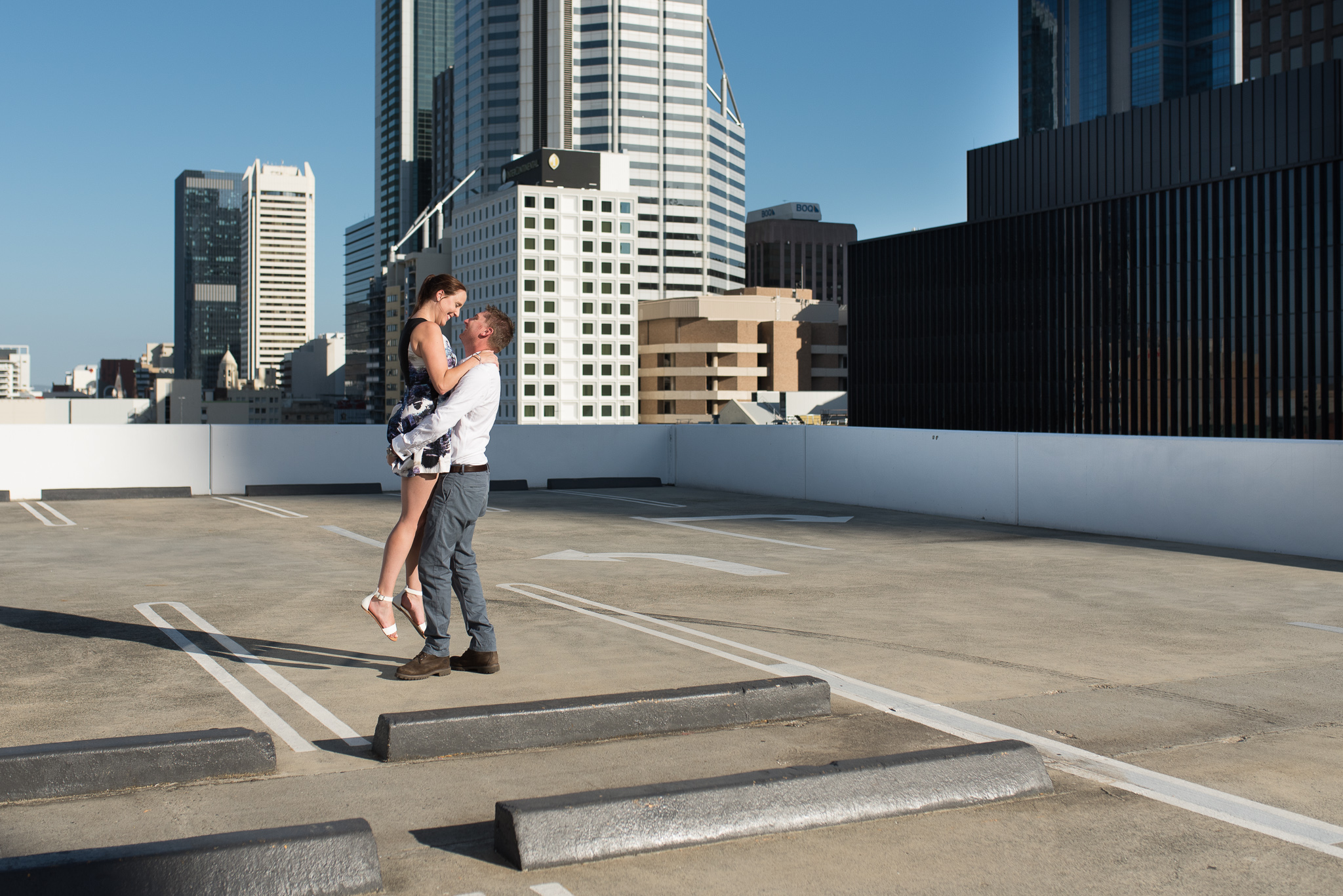 Boy lifts his fiancee up on carpark rooftop with Perth cityscape behind