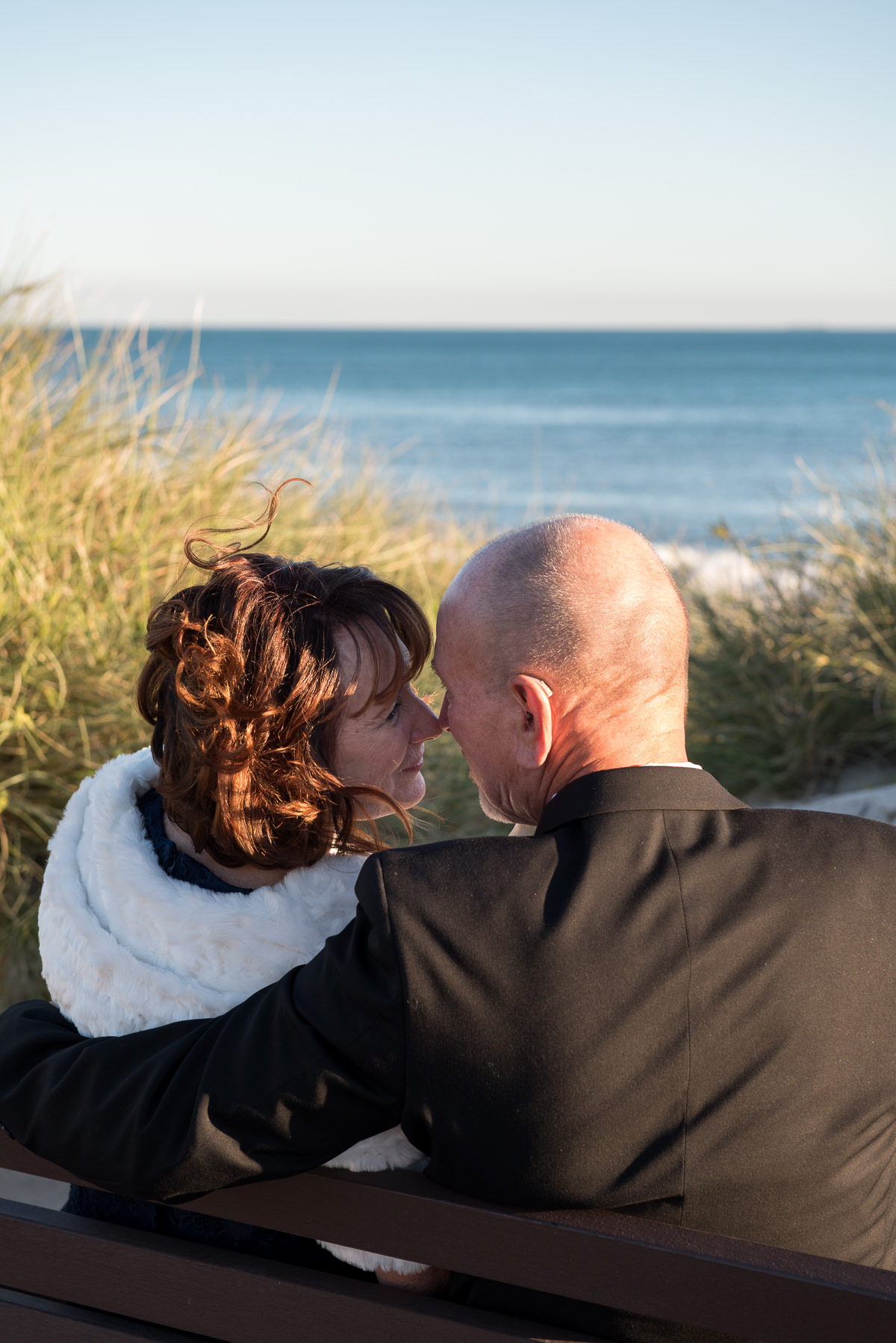 Older wedding couple sitting on bench touching noses next to the ocean