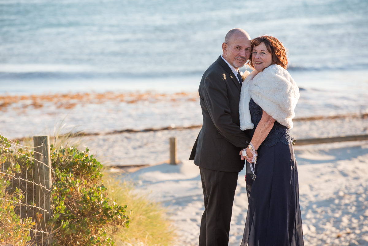 Older wedding couple with the ocean behind them standing cheek to cheek