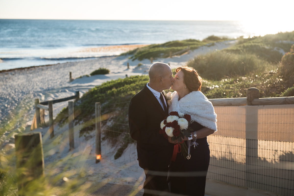 Older wedding couple kissing with the beach behind them, bride holding bouquet and wearing a white wrap