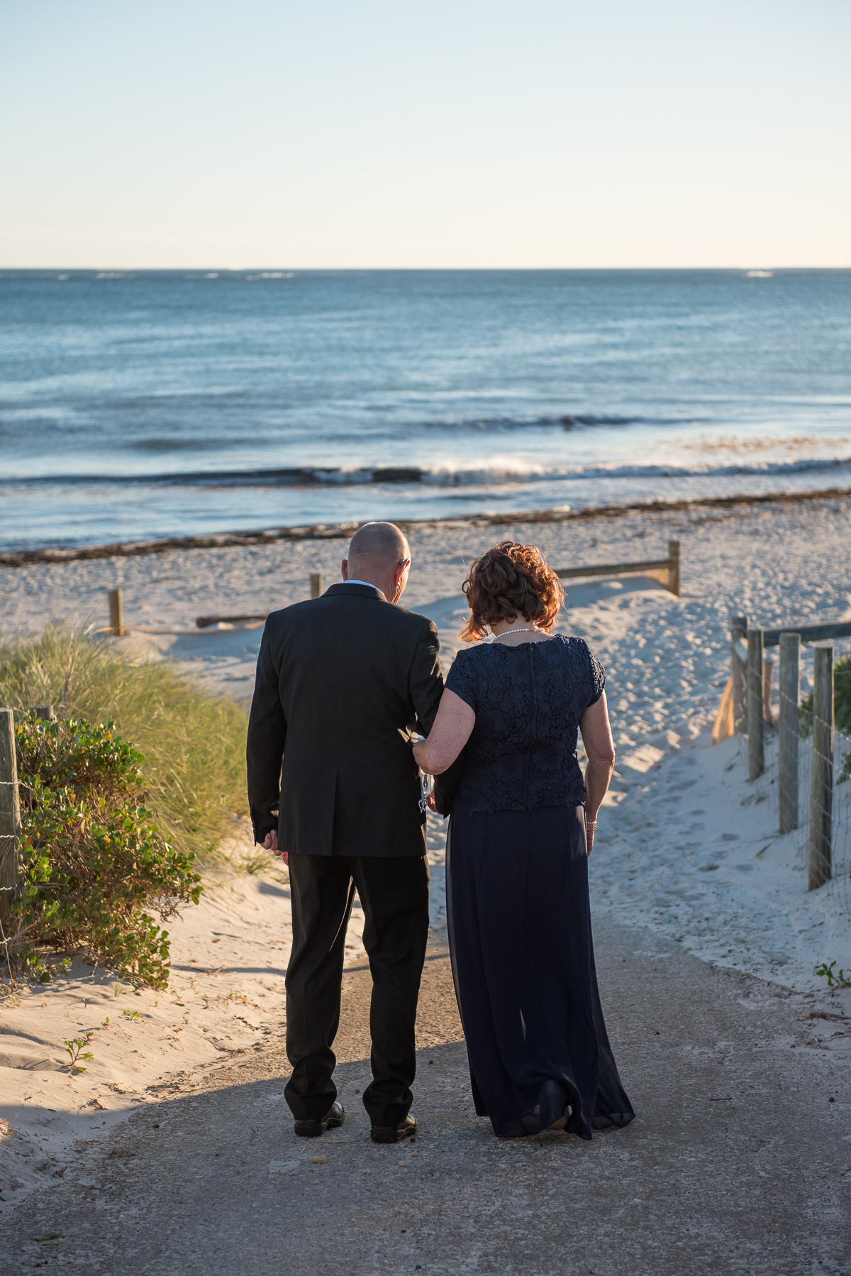 Older wedding couple walking down path towards the beach at Mindarie