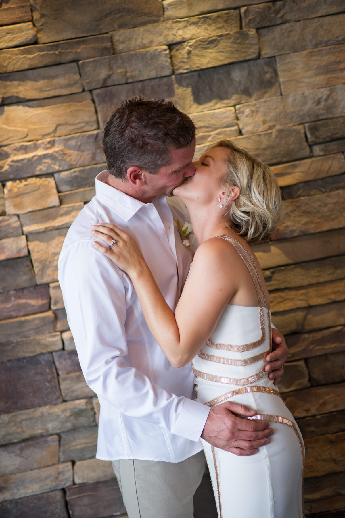 Bride and groom standing against brick wall at The Breakwater kissing